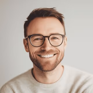 Smiling man with short brown hair, beard, and glasses wearing a light-colored sweater against a plain background.